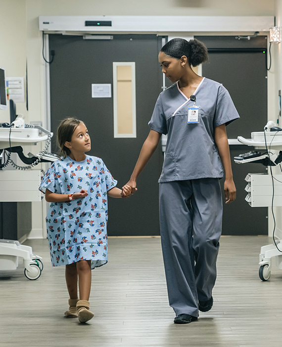 Nurse and little girl in a hospital gown walk down a hospital hallway holding hands.
