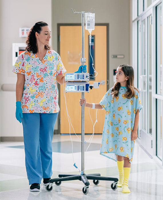 A nurse and a little girl holding on to an infusion pole with 2 infusion pumps attached walk down a hospital corridor.