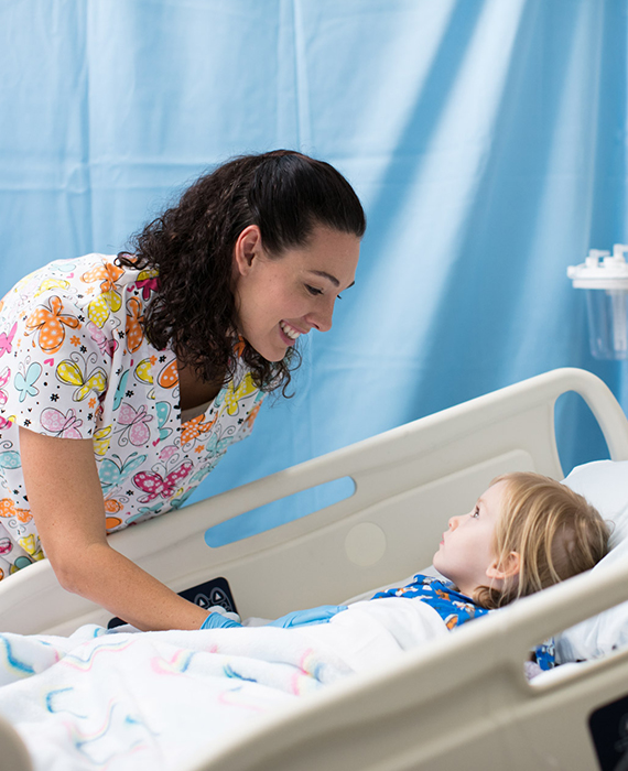 A female nurse at the beside of a pediatrics patient who is laying in a hospital bed in a hospital room.