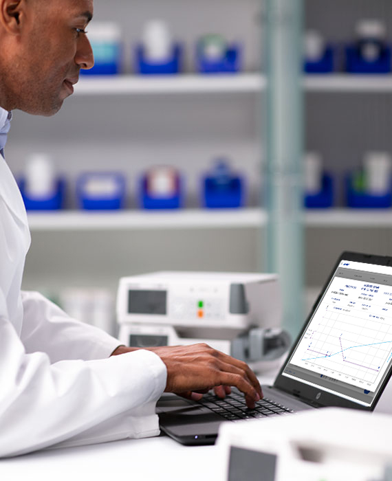 Pharmacist sitting at a desk reviewing data on a laptop. There are several infusion pumps on the table next to him.